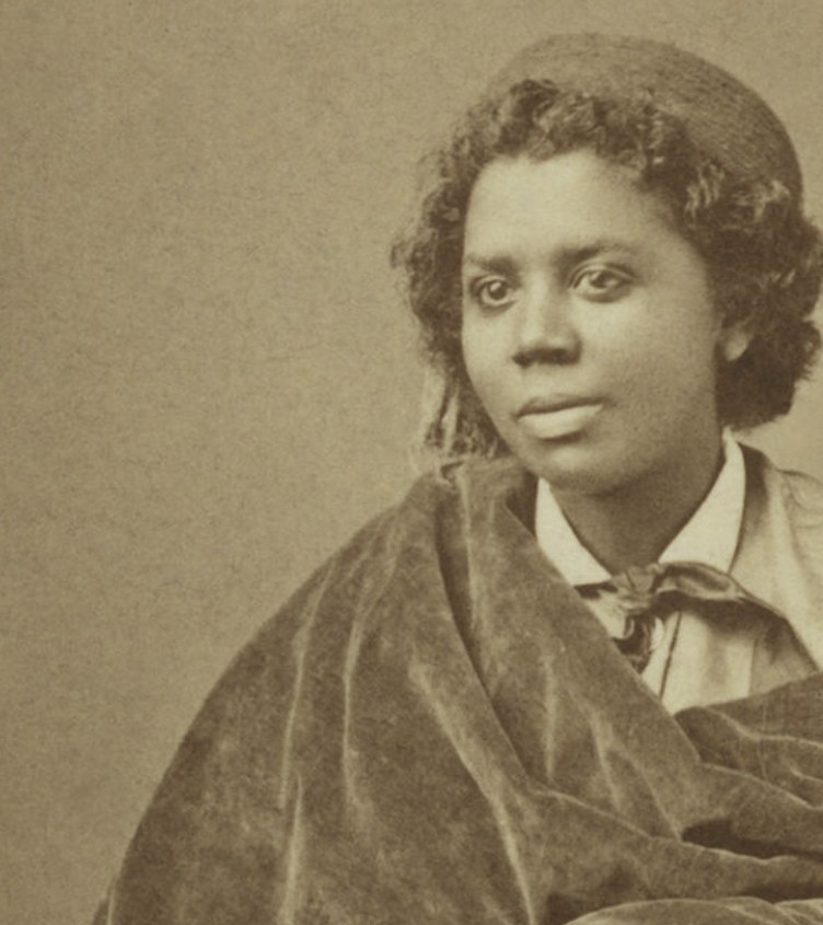 Historic studio portrait photograph of Edmonia Lewis, wearing a velvet cloak over a collared blouse with a bow